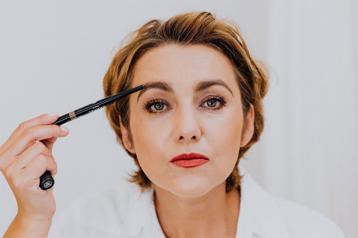 woman with red lipstick applying makeup to her eyebrows