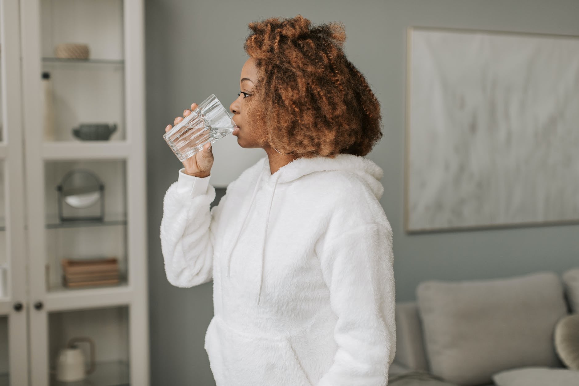 A woman stands in a comfortable sweater as she sips a glass of water, taking care of her well-being