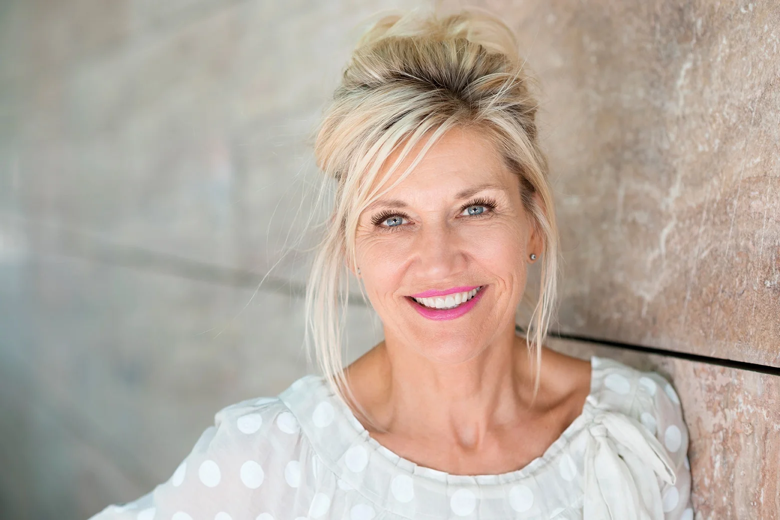 middle-aged blonde woman smiling and wearing pink lipstick and lash extensions