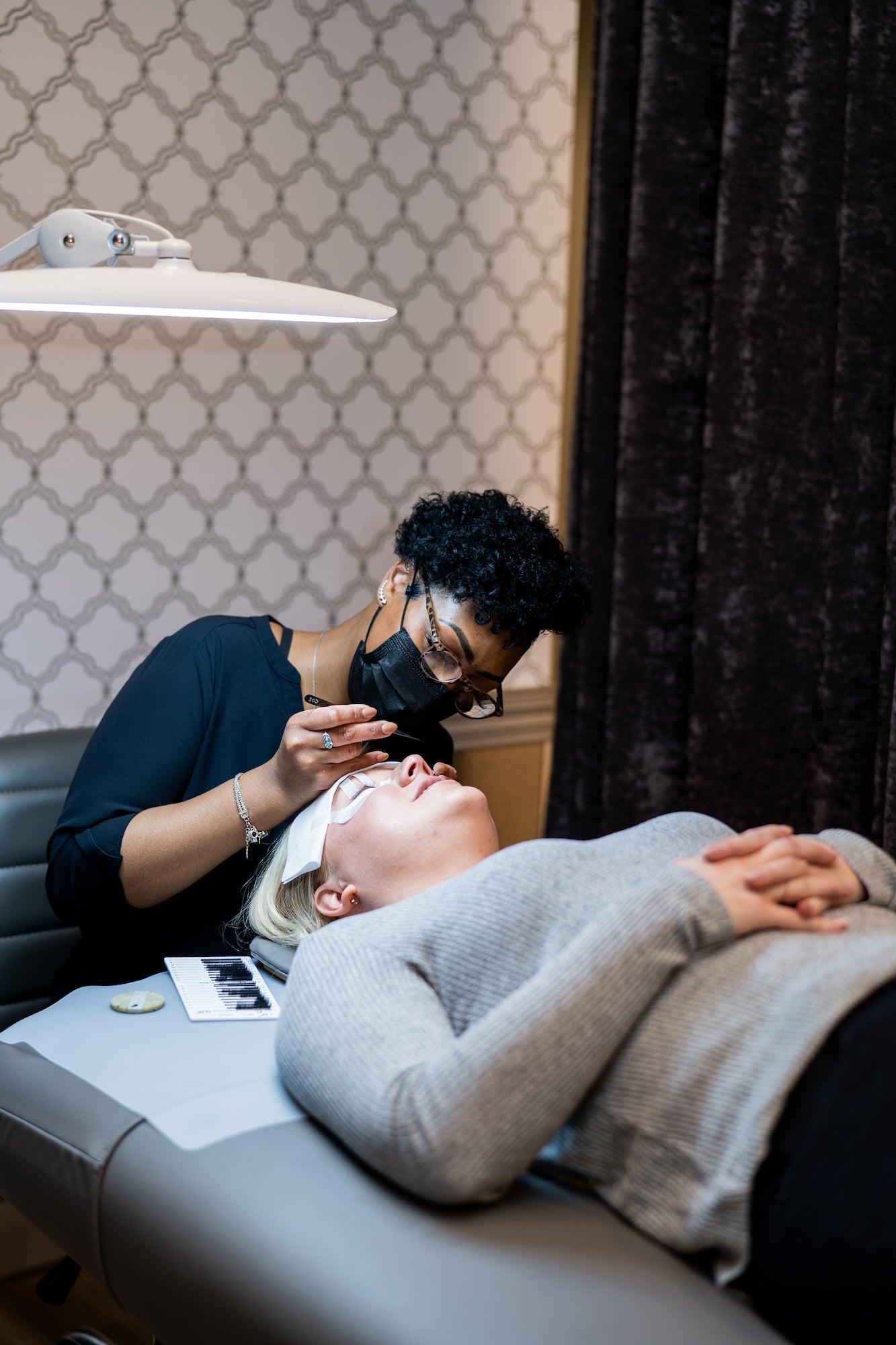 a lash stylist sitting while apply lash extensions to her relaxing guest