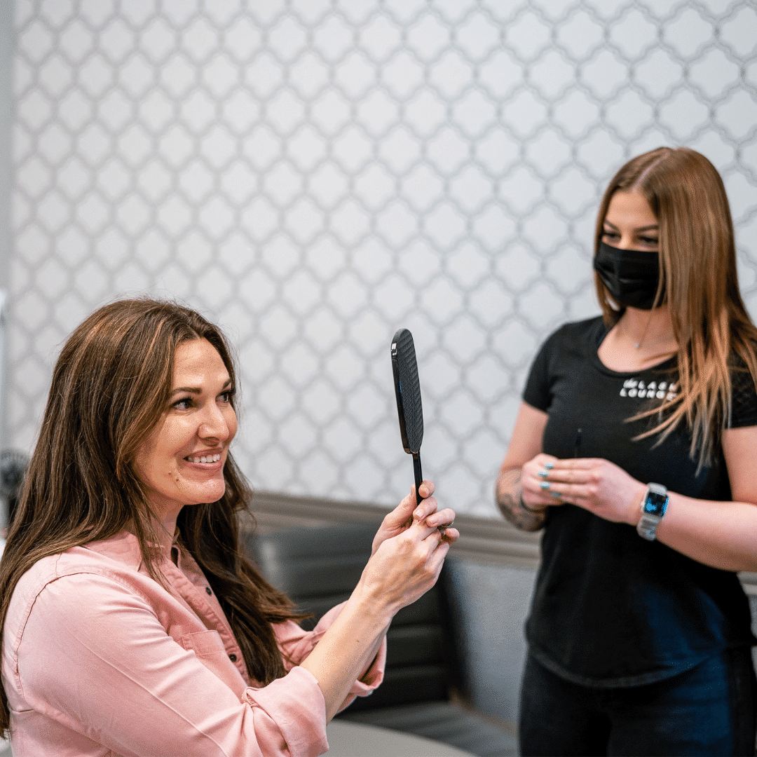 Lash Lounge guest looking at her new lash extensions with a handheld mirror while her lash stylist stands to the side.