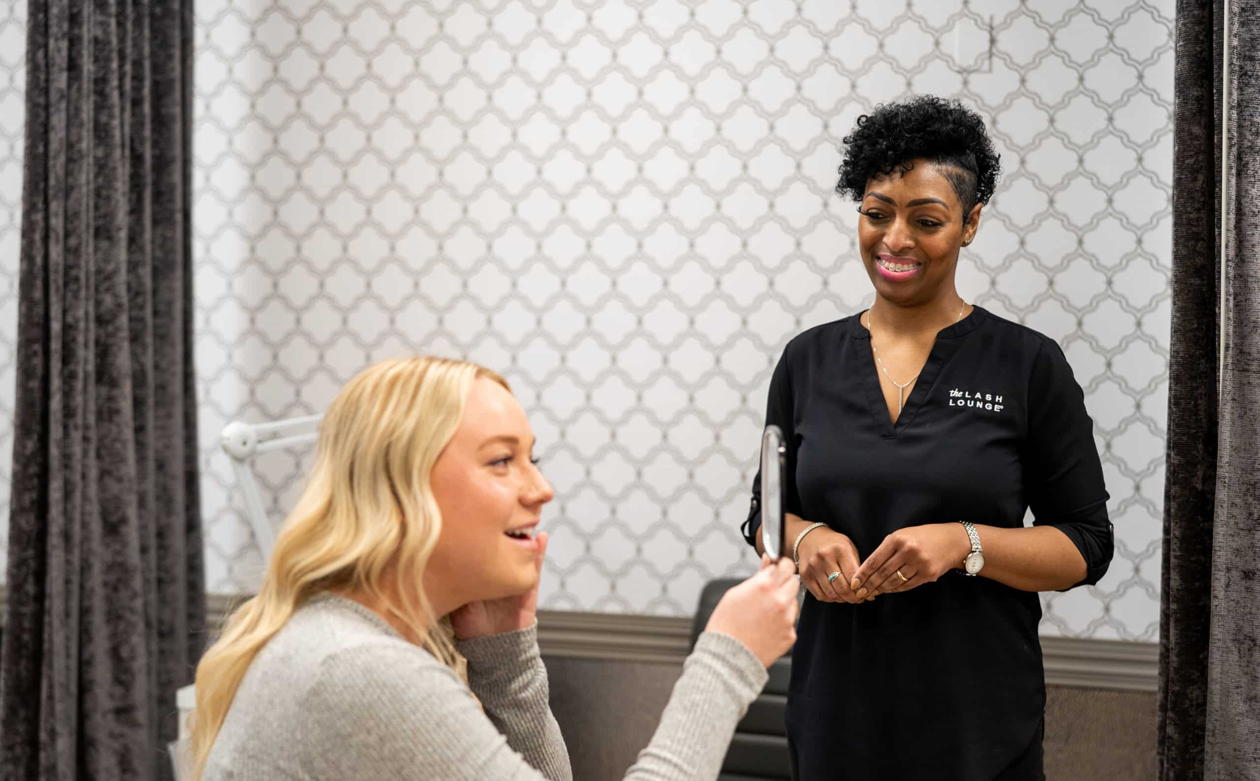 An lash technician from The Lash Lounge smiles as a guest admires her new lashes