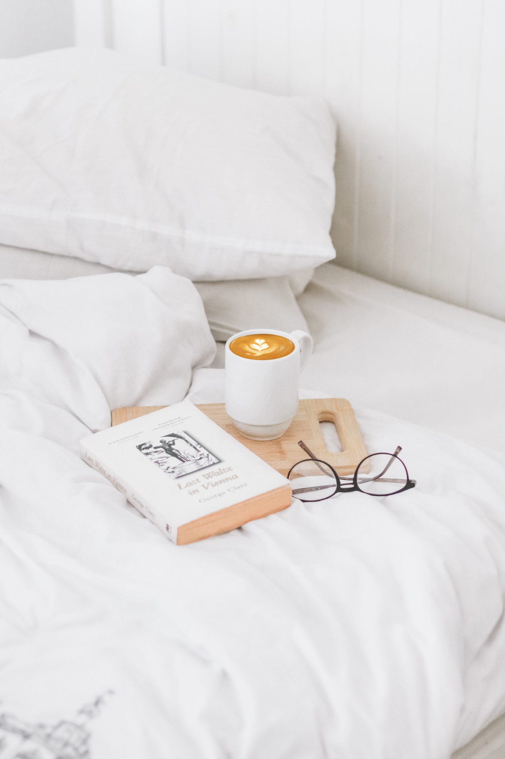 Reading glasses, a book, and a cup of coffee, are placed on a bed, and are ideas for investing in yourself on a relaxing day
