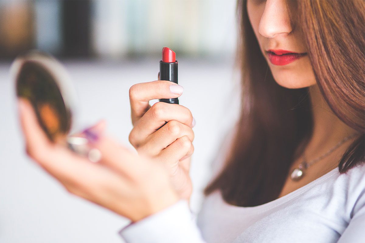 Half of a woman's face shows as she holds a lipstick and handheld mirror to see her new makeup look