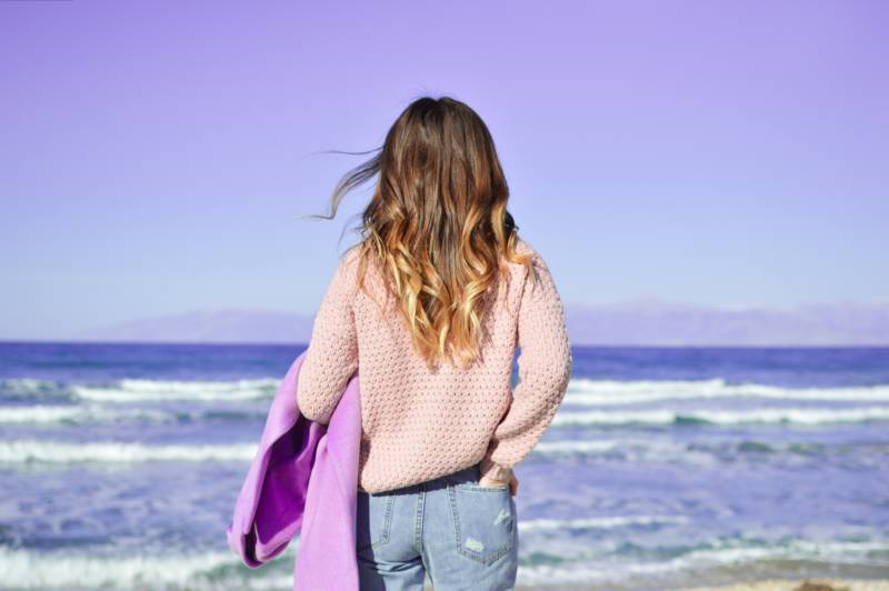 Girl from behind with curly hair holding a purple jacket looks at the ocean and sky