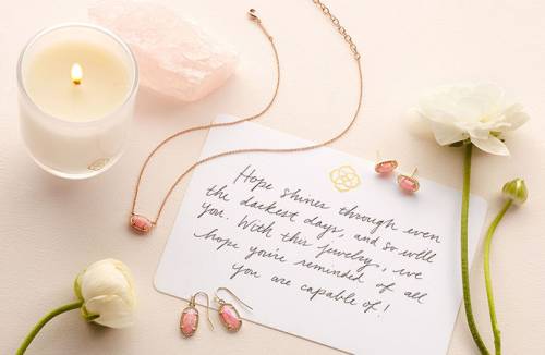 an array of items placed on a flat surface, including: Kendra Scott jewelry, flowers, a candle and a note