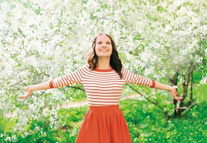 woman in striped dress opens her arms and smiles with trees and flowers in the back