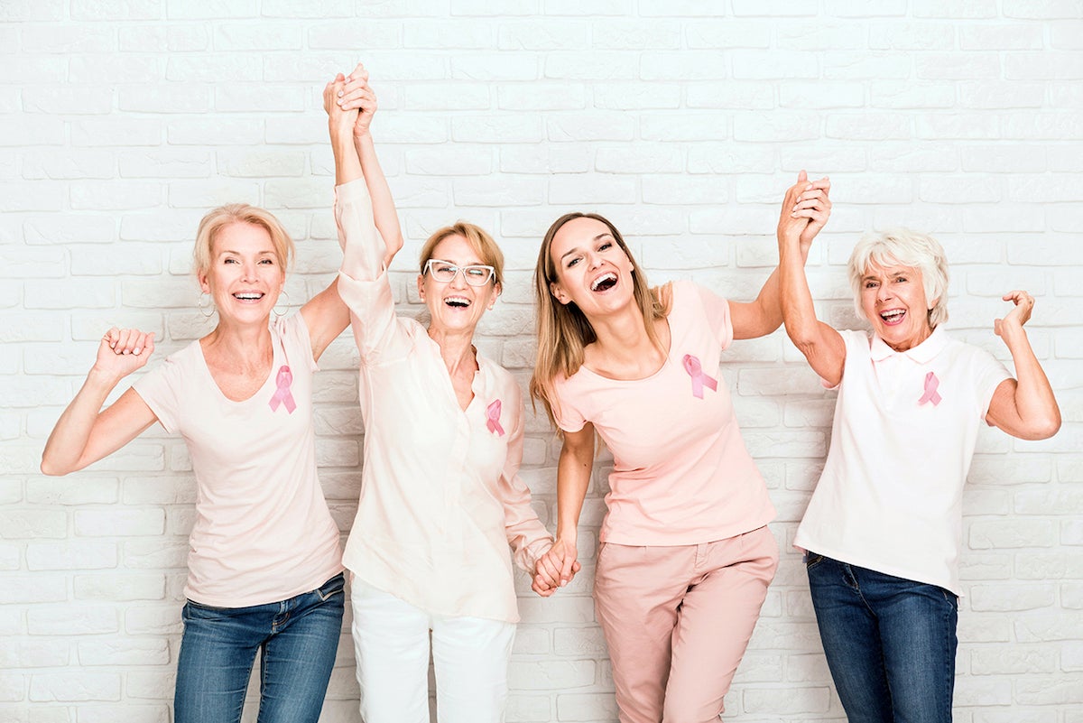 A group of women smiling and holding hands wearing breast cancer symbols in front of brick wall