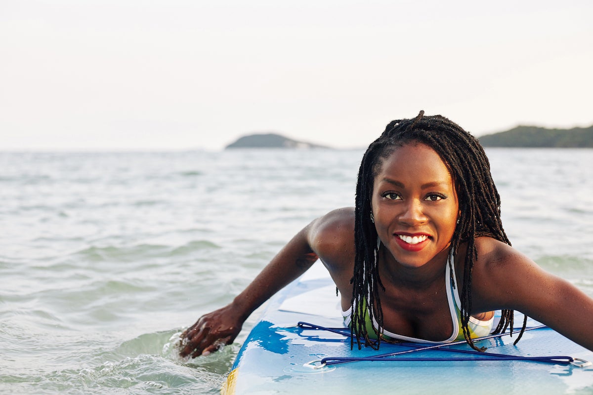 black woman in ocean swimming on surf board and smiling with eyelash extensions