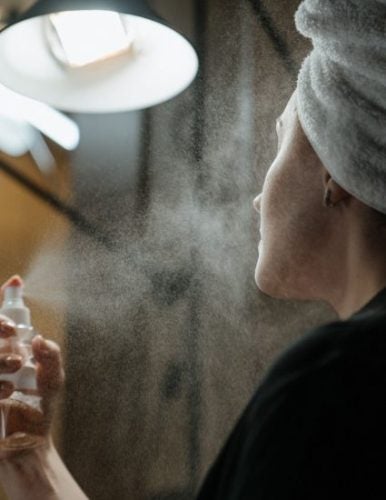 woman with towel on her hair spritzing her face with a spray