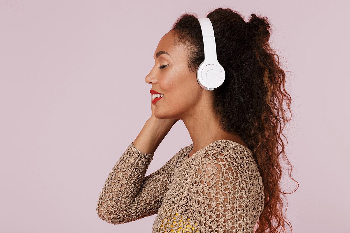 A young black woman smiling with her eyes closed listening to music with white headphones on her ears.