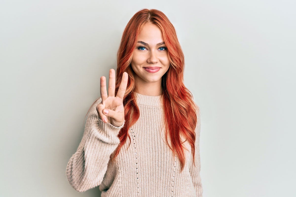 A woman with red hair and eyelash extensions smiles and holds up three fingers