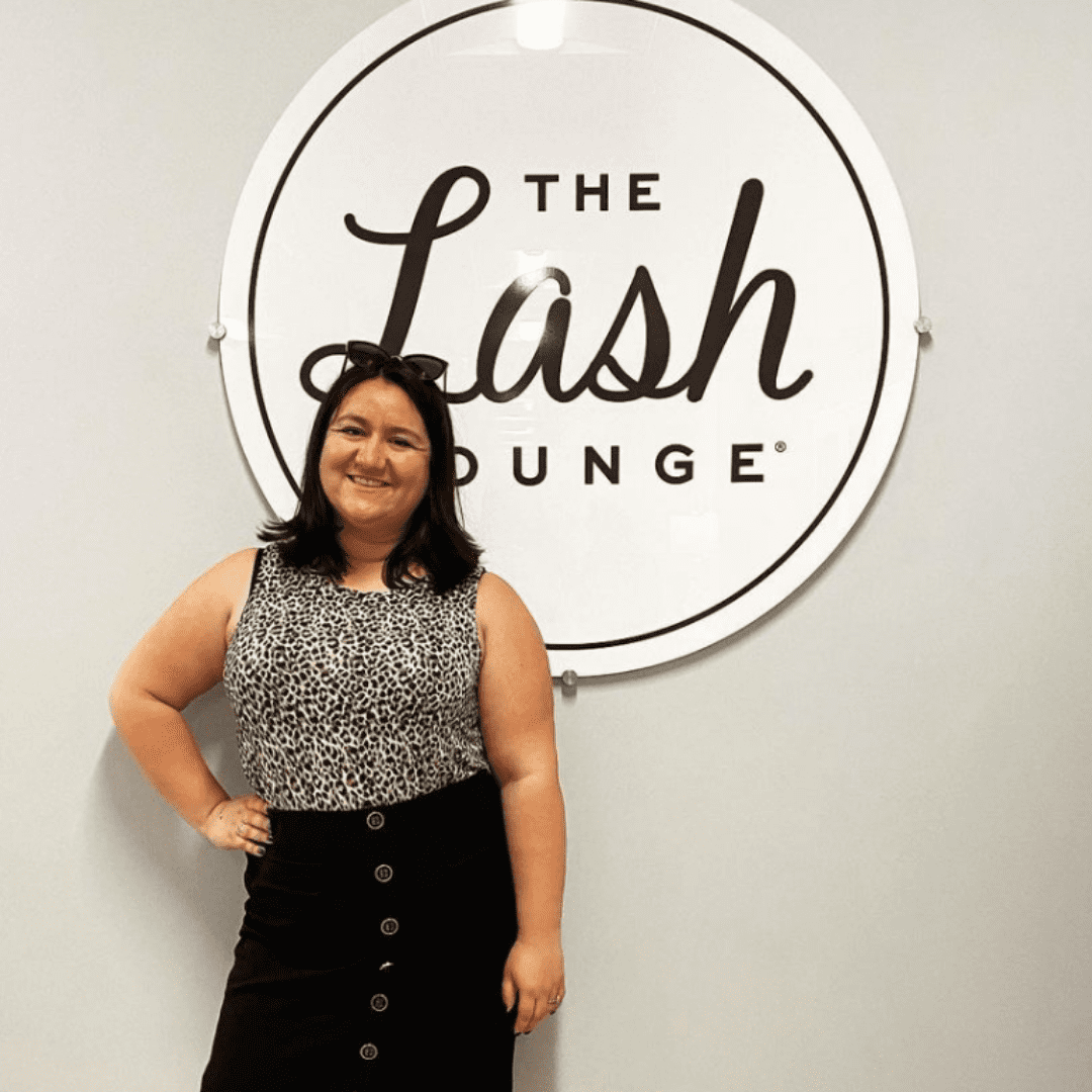 Young brunette woman and employee of The Lash Lounge standing in front of the logo