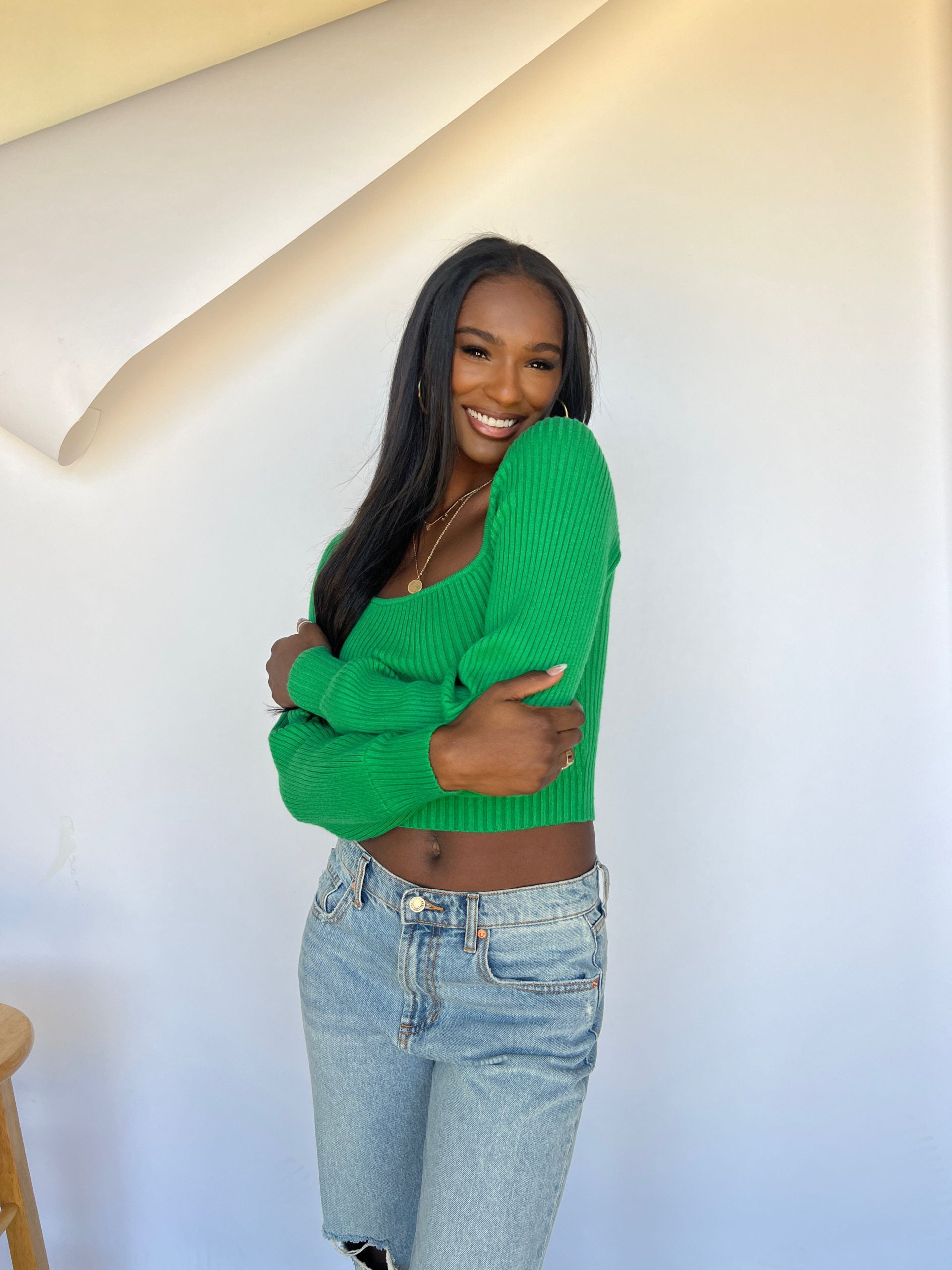 Lifestyle influencer and Lash Lounge guest, Nikki Baize, posing in front of a white backdrop while smiling and wearing a green sweater and jeans.