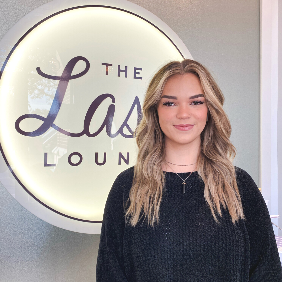 Lash stylist, Kassidy, smiling in front of The Lash Lounge light up sign.