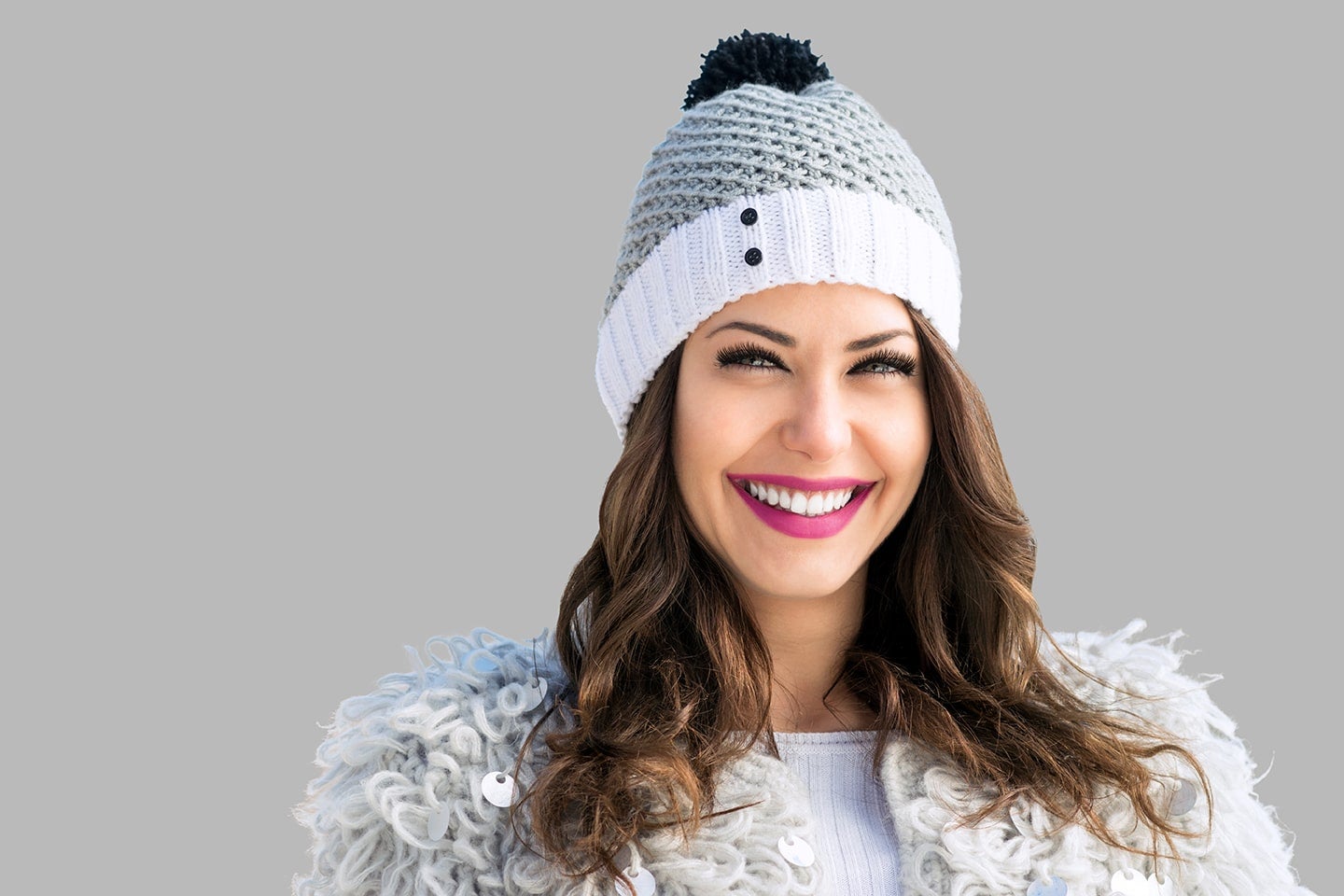 A woman smiles while wearing a winter hat and holiday makeup with a red lip and lashes