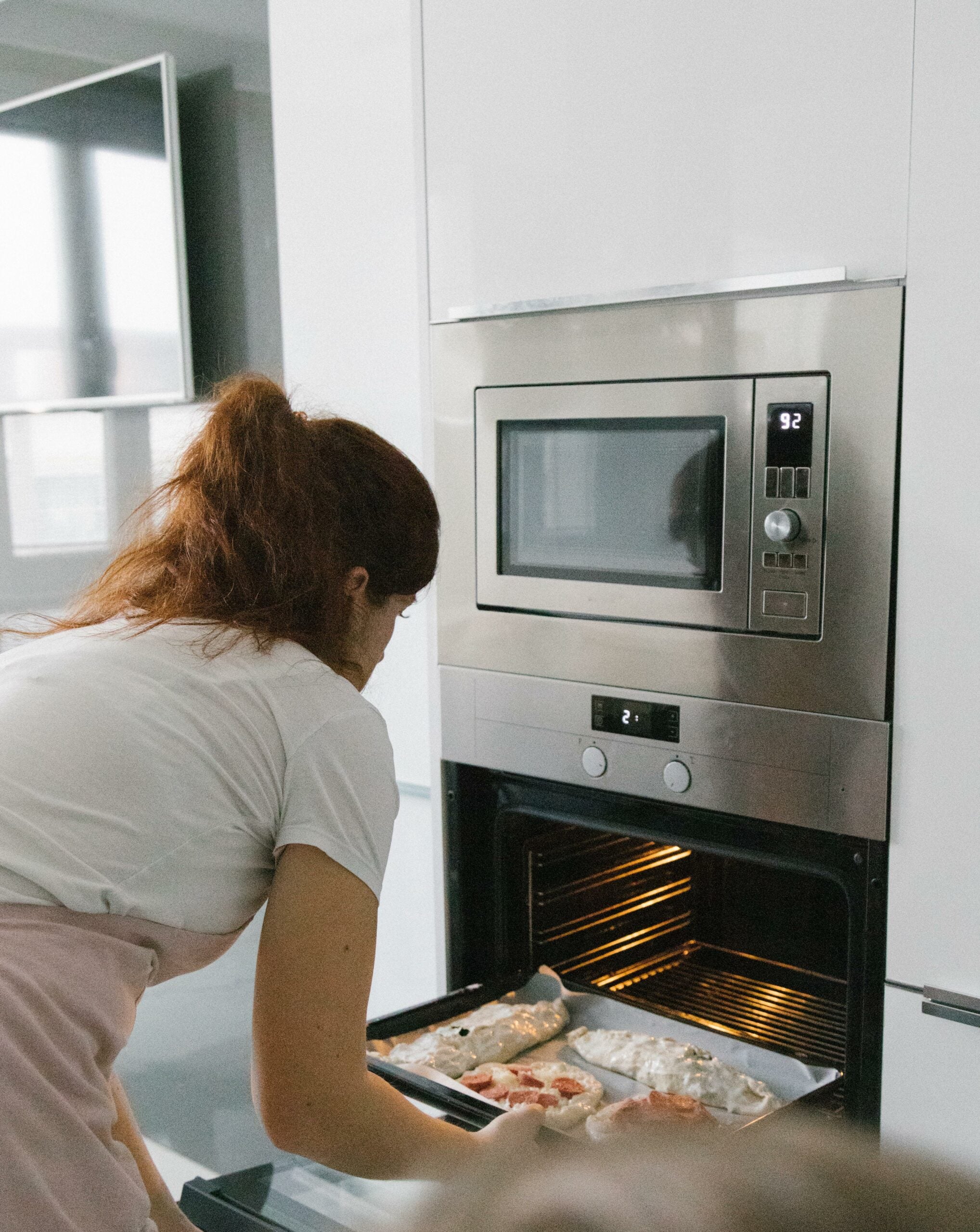 A woman bending down and putting pizza on a pan into an oven